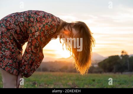 Lächelnde junge Frau mit braunen Haaren, die sich während des Sonnenuntergangs verbiegen Stockfoto