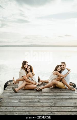 Töchter umarmen Mutter und Vater, während sie auf dem Steg sitzen see gegen Himmel Stockfoto