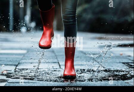 Mädchen tragen Gummistiefel spritzt Wasser beim Springen in Pfütze Auf Fußweg Stockfoto