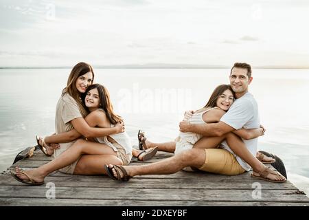 Töchter umarmen Eltern, während sie auf dem Steg am See sitzen Himmel Stockfoto