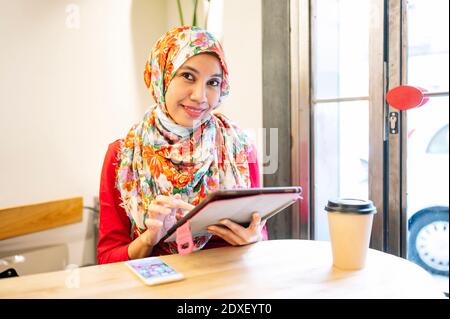 Lächelnde Frau trägt Hijab hält digitale Tablette im Café sitzen Stockfoto