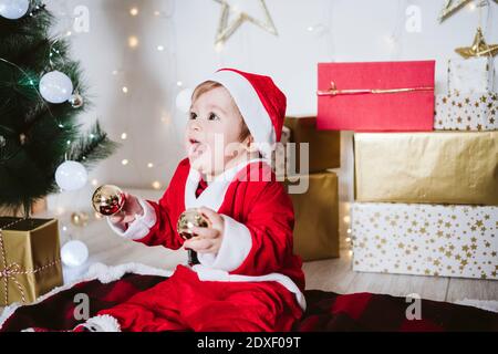 Verspielter Junge im Weihnachtsmann Kostüm spielt mit Kugel Während der Weihnachtszeit auf der Decke zu Hause sitzen Stockfoto