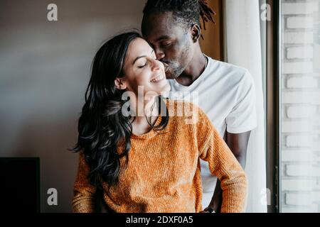 Junger Mann küsst glücklich schwangere Frau durch Fenster zu Hause Stockfoto