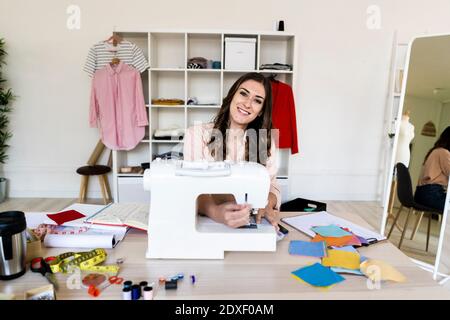 Junge Schneiderin lächelnd, während sie mit Nähmaschine sitzt studio Stockfoto