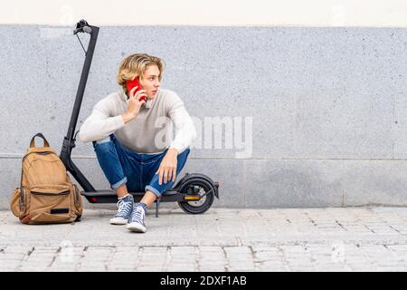 Mann mit Rucksack, der auf dem Mobiltelefon spricht, während er auf dem Gerät sitzt Elektroroller gegen die Wand schieben Stockfoto