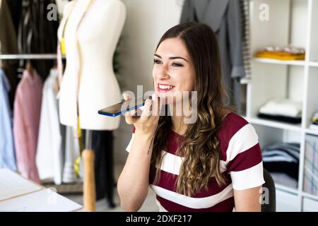 Lächelndes weibliches Design professionelle Senden Voicemail über Smartphone während Sitzen im Studio Stockfoto