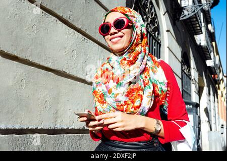 Lächelnde muslimische Frau hält Smartphone, während sie durch das Gebäude geht An sonnigen Tag Stockfoto