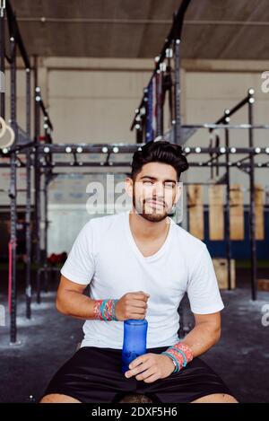 Lächelnder Mann hält Wasserflasche, während er im Fitnessstudio sitzt Stockfoto