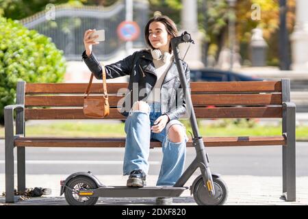 Lächelnde Frau, die Selfie über das Smartphone nimmt, während sie darauf sitzt Bank von Elektro-Push-Roller Stockfoto