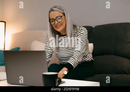 Lächelnde ältere Frau mit Laptop, während sie auf dem Sofa sitzt Zu Hause Stockfoto