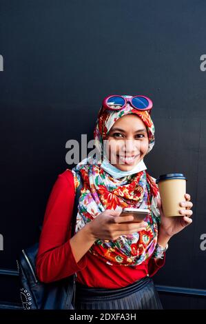 Lächelnde Frau trägt Hijab hält Handy und Kaffeetasse Gegen schwarze Wand während Pandemie Stockfoto