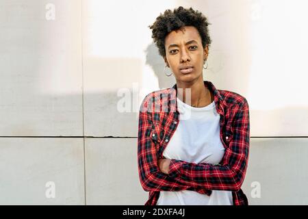 Selbstbewusste junge Afro-Frau, die mit gekreuzten Armen gegen Weiß steht Wand Stockfoto