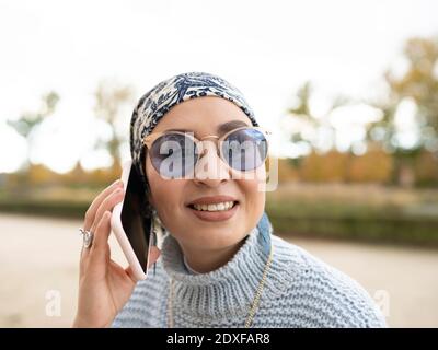 Junge Frau mit Kopftuch und Sonnenbrille lächelnd während des Gesprächs Mobiltelefon im Freien Stockfoto