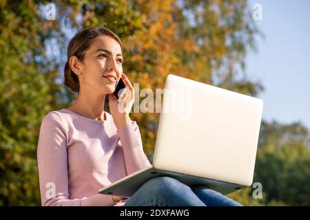 Junge Unternehmerin im Gespräch auf dem Smartphone, während sie mit Laptop im Park im Herbst Stockfoto