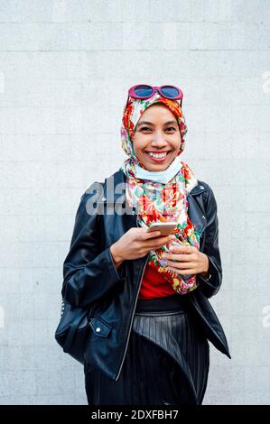 Lächelnde Frau in Lederjacke, während sie das Handy dagegen hält Wand während COVID-19 Stockfoto