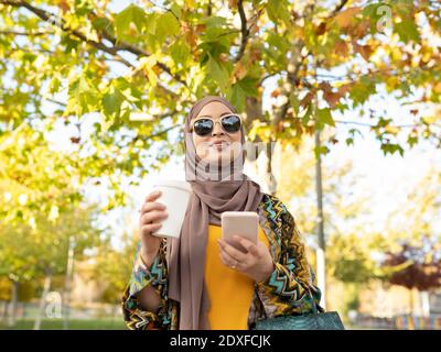 Junge Frau trägt Hijab und Sonnenbrille hält Kaffee Tasse während Im Freien stehen Stockfoto