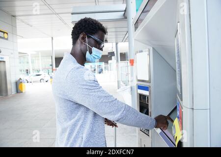 Reifer männlicher Profi mit Fahrkartenautomaten am Bahnhof während Coronavirus Stockfoto
