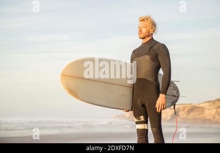 Blonde Mann trägt Neoprenanzug trägt Surfbrett beim Blick weg auf Strand gegen Himmel Stockfoto