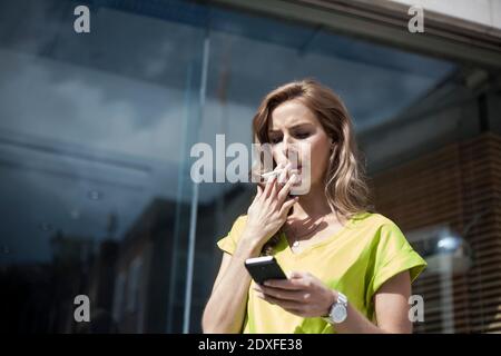 Junge Unternehmerin mit Smartphone beim Rauchen gegen Büro Gebäude an sonnigen Tag Stockfoto