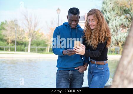 Afrikanischer schwarzer Mann und Rotschopf kaukasische Frau lächeln und Blick auf ihr Smartphone in einem Park. Junge multirassische Paar Porträt. Stockfoto