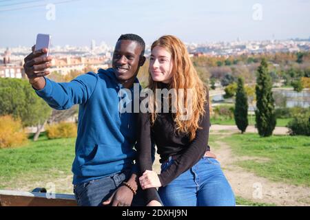 Afrikanischer schwarzer Mann und rothaarige kaukasische Frau lächeln und machen ein Selfie mit ihrem Smartphone in einem Park. Junge multirassische Paar Porträt. Stockfoto