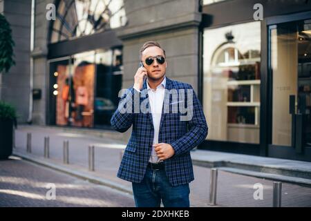 Junger stilvoller Mann in Sonnenbrille im Gespräch auf einem Handy in der Innenstadt. Trägt eine blaue Jacke und weißes Hemd. Stockfoto