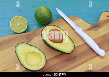 Top-Gericht mit einer geschnittenen Avocado, Limetten und einem Messer auf Holzplatte und blauem Hintergrund. Stockfoto