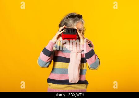 Ältere Frau in legerer Kleidung, die ein Foto mit einem sofortige Kamera vor gelbem Hintergrund Stockfoto
