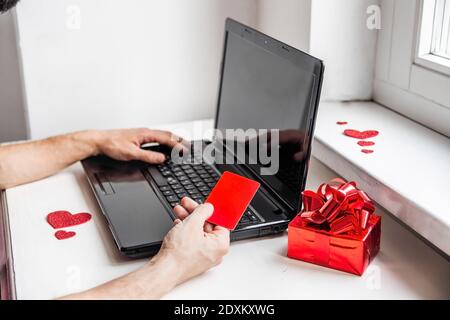 Valentinstag Online-Shopping-Konzept. Nahaufnahme der Hände eines Mannes Halten Sie eine rote Nachstellung von Kreditkarte und Geschenk in der Nähe eines Laptops. Stockfoto