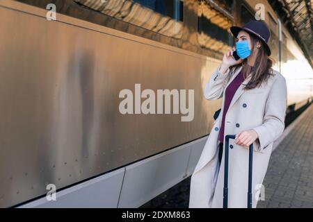 Frau in Maske spricht im Stehen am Telefon In der Nähe von Personenzug Auto am Bahnhof Stockfoto