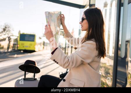 Junge Frau sitzt an der Bushaltestelle nad hält Stadtplan. Mädchen schaut auf die Papierkarte und sucht nach dem Ort, den sie braucht. Reisekonzept Stockfoto