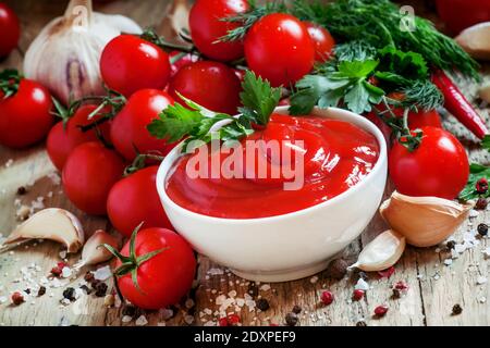 Tomatenketchup-Sauce mit Knoblauch, Gewürzen und Kräutern mit Kirschtomaten in einer weißen Porzellanschüssel auf altem Holztisch, selektiver Fokus Stockfoto