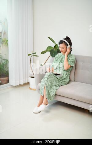 magier der fokussierten asiatischen Frau mit Kopfhörer und schreiben Notizen, während Sie zu Hause auf dem Sofa sitzen Stockfoto