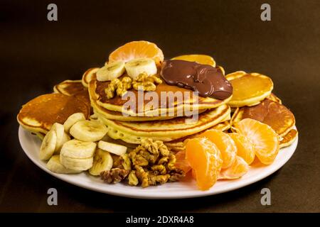 Kleine und große, gut gebackene Pfannkuchen, die von einem weißen flachen Teller mit Schokolade Haselnuss-Sahne, Walnuss, Mandarine und Banane serviert werden Stockfoto