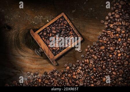 Nahaufnahme von gerösteten Kaffeebohnen in einer kleinen Holzschublade einer Kaffeemühle auf einem Holztisch mit vielen Kaffeebohnen. Stockfoto