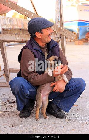 Ein Schiffsreparaturarbeiter spielt mit einem Straßenhund (Welpe) in der Nähe eines alten roten Holzboots in der Werft. Es gibt Holz und Reparaturgeräte in der Nähe Stockfoto