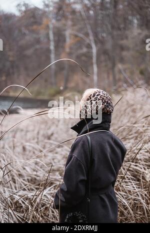 Frau im Leopardendruck Kopftuch steht zwischen dem trockenen Gras und schaut auf den Wald und den See. Hochwertige Fotos Stockfoto