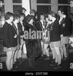 1960er Jahre, historisches, großes Lächeln als Hockeyteam der Schulmädchen in ihren matschigen Stiefeln und Blazern wird mit der Siegertrophäe von einer Dame im Pelzmantel aus Fife, Schottland, Großbritannien, ausgezeichnet. Stockfoto