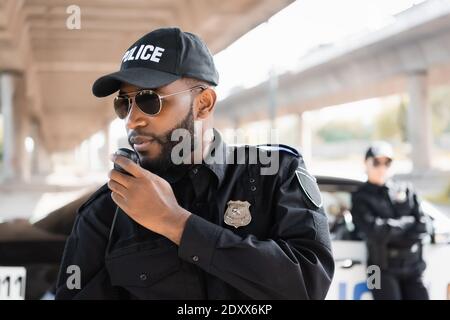 afroamerikanischer Polizist, der auf dem Radiogerät in der Nähe der Polizistin spricht Bei unscharfem Hintergrund im Freien Stockfoto