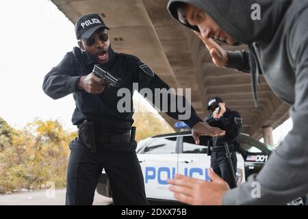 Wütender afroamerikanischer Polizist, der mit Pistole auf verschwommenes ergeben zielt Straftäter im Vordergrund auf der städtischen Straße Stockfoto