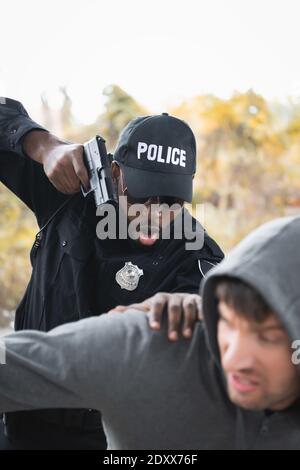 Aggressiver afroamerikanischer Polizist, der mit Pistole anvisiert, während er eine Kapuze festnahm Straftäter auf verschwommenem Vordergrund im Freien Stockfoto