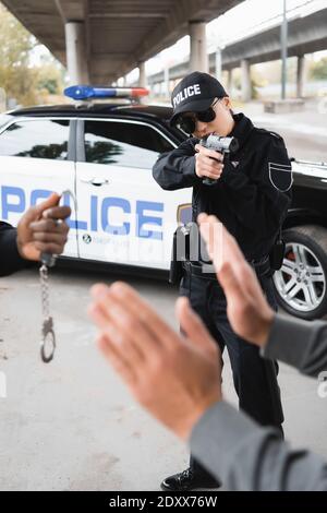 Polizistin mit Pistole auf übergebenen Täter mit verschwommenem afrikaner zielen amerikanischer Kollege im Vordergrund draußen Stockfoto