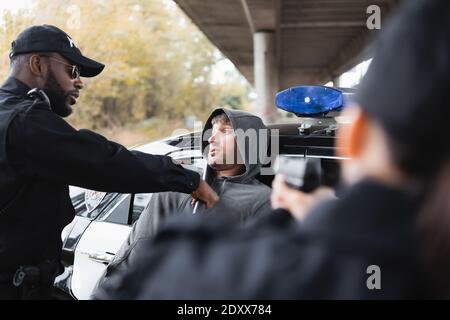 afroamerikanischer Polizist mit Knüppel verhaften Kapuze Täter mit verschwommen Kollege mit Waffe im Vordergrund im Freien zielen Stockfoto