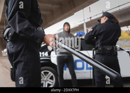 afroamerikanischer Polizist hält Knüppel in der Nähe von Kollegen mit dem Ziel Pistole und Straftäter auf unscharfem Hintergrund im Freien Stockfoto