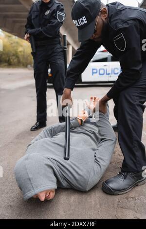 afroamerikanischer Polizeibeamter mit Knüppel, der einen vermummten Täter festnahm, liegt Auf der Straße auf unscharfem Hintergrund Stockfoto
