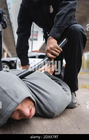 afroamerikanischer Polizist mit einem Knüppel, der einen vermummten Täter festnahm, auf dem er lag Straße auf unscharfem Hintergrund Stockfoto