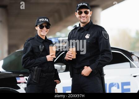 Lächelnde Polizisten in Sonnenbrillen halten Kaffee zum Mitnehmen in der Nähe des Autos Bei unscharfem Hintergrund im Freien Stockfoto