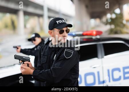 Polizist in Sonnenbrille hält Gewehr und schaut weg in der Nähe von Kollegen Und Auto auf verschwommenem Hintergrund Stockfoto