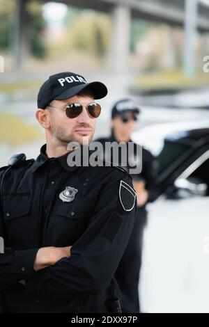 Polizist in Sonnenbrille stehend mit gekreuzten Armen in der Nähe von Kollegen und automatisch bei unscharfem Hintergrund Stockfoto