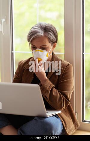 Grauhaarige Freiberuflerin, die mit einem Laptop arbeitet und eine medizinische Maske auf ihrem Gesicht trägt, während sie auf einer Fensterbank sitzt. Warmfarbiges Bild. Geschäftskonzept
